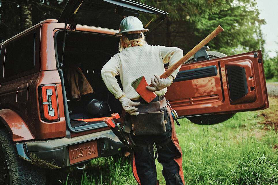 A person in work gear stands at the open back of a red SUV, holding an axe and facing away, with trees and grass in the background