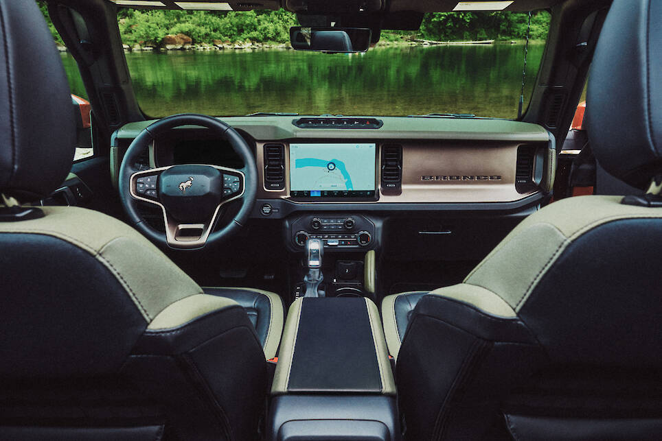 View of the interior of a Ford Bronco, showing the dashboard, steering wheel, touchscreen display, and front seats with a lake and trees visible through the windshield