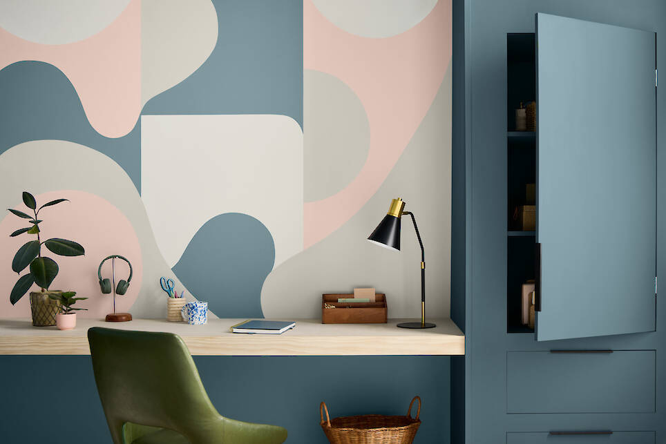 A modern workspace with a green chair, desk lamp, potted plant, stationery, and abstract pastel wall art inspired by the Valspar 2027 Color of the Year. Blue cabinets are partia