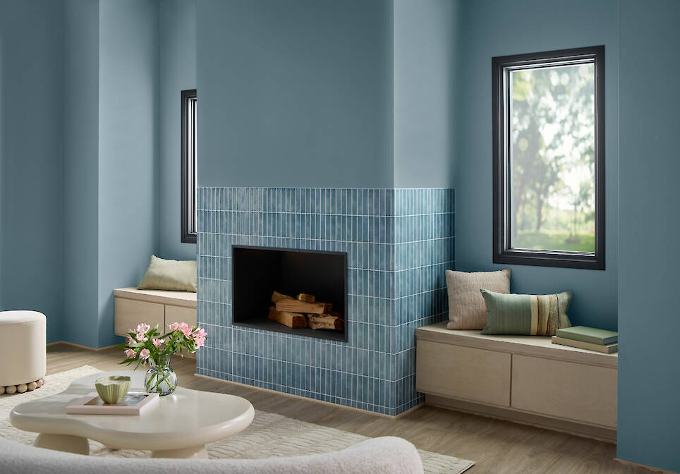 A living room with a blue-tiled fireplace, built-in window seats with cushions, blue walls painted in Valspar 2027 Color of the Year, wood flooring, and a round coffee table wit