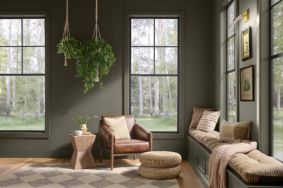 A cozy reading nook with a leather chair, poufs, window bench, hanging plant, and large windows overlooking a green forest is beautifully accented by touches of Behr 2027 Color
