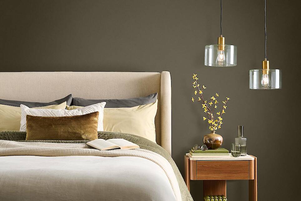 Modern bedroom with a beige upholstered bed, neutral bedding, wooden nightstand with a vase of yellow flowers, book, and two hanging glass pendant lights—all beautifully accen