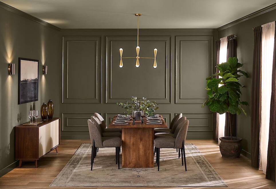 A modern dining room with a wooden table, eight chairs, a sideboard, large potted plant, and gold chandelier, featuring green walls painted in Behr 2027 Color of the Year and na