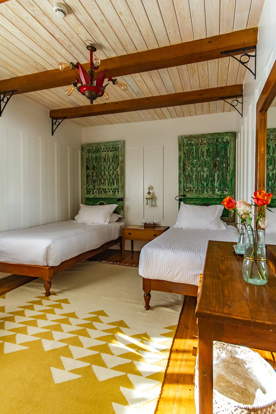 A bedroom with two single beds, a wooden ceiling, green wall art, a yellow patterned rug, and a wooden dresser with vases of red and orange flowers