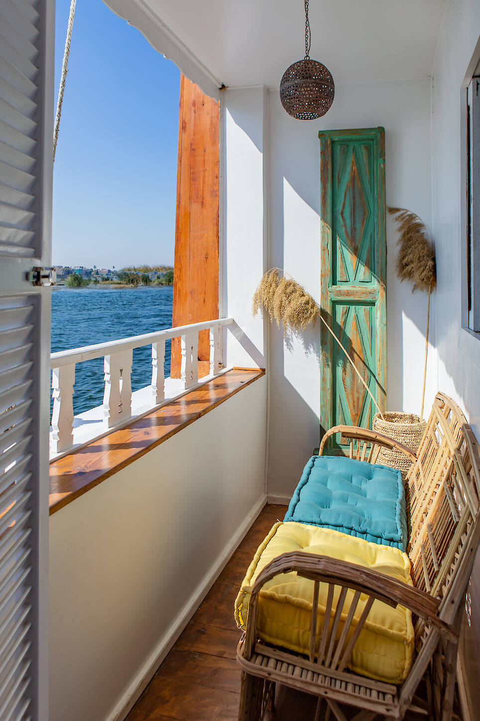 A narrow balcony with wicker chairs, colorful cushions, a wooden railing, dried grass decor, a green rustic door, and a view of water under a clear blue sky