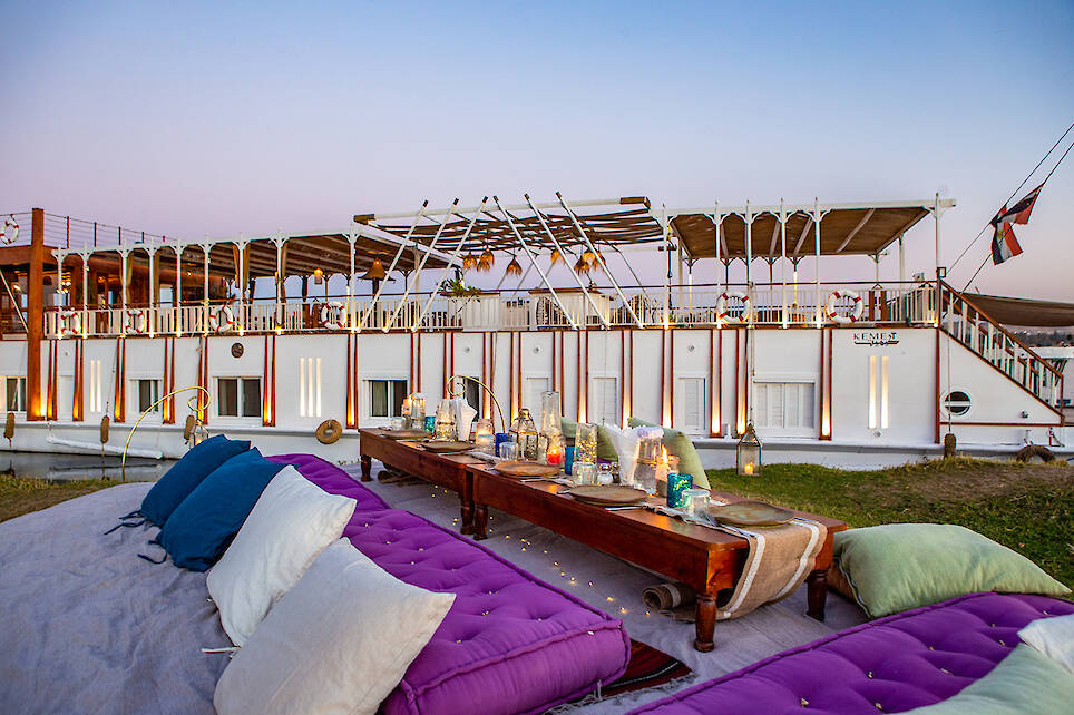 Outdoor picnic setup with colorful cushions and a low wooden table arranged on a lawn next to a docked riverboat at sunset