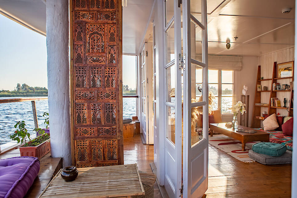 Sunlit houseboat interior with wooden floors, large windows, ornate carved panel, cushions, low table, and plants. Water and shoreline visible through windows and open doors