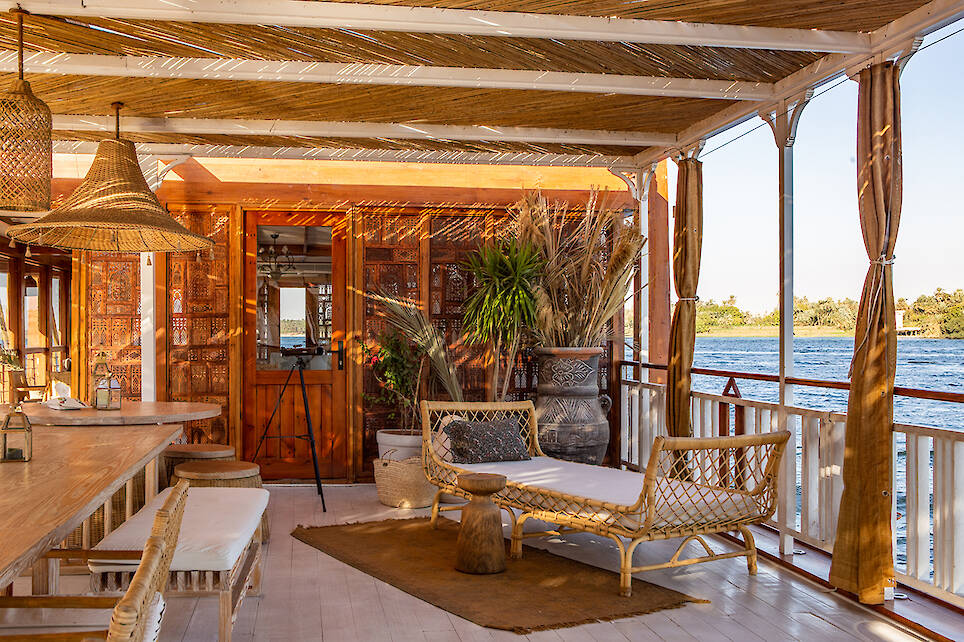 Sunlit boat deck with wicker furniture, potted plants, wooden decor, and river view; shaded by a bamboo roof