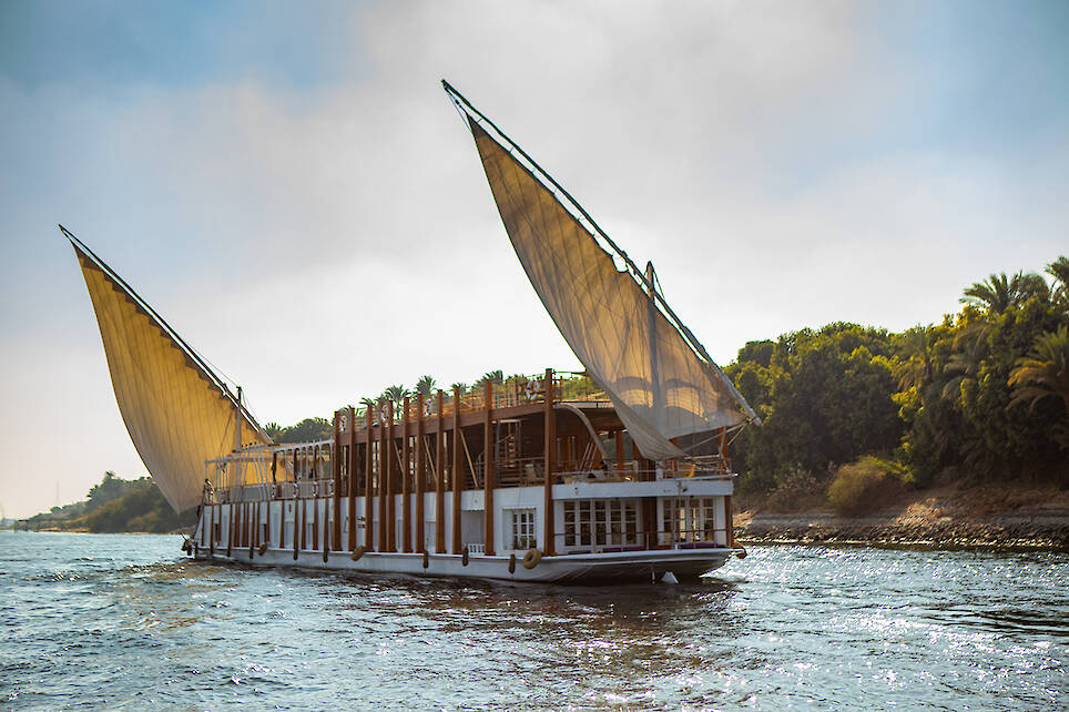 A large traditional sailboat with two triangular sails moves along a river, with green trees lining the shore in the background under a partly cloudy sky