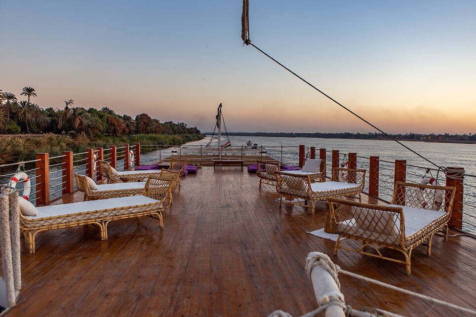 Open deck of a riverboat with wicker lounge chairs facing the water, wooden flooring, and railings, set against a calm river and a sunset sky