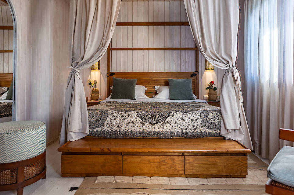 A neatly made bed with patterned bedding, green pillows, and beige curtains on a wooden frame, set in a bright bedroom with light walls and soft natural light from a window
