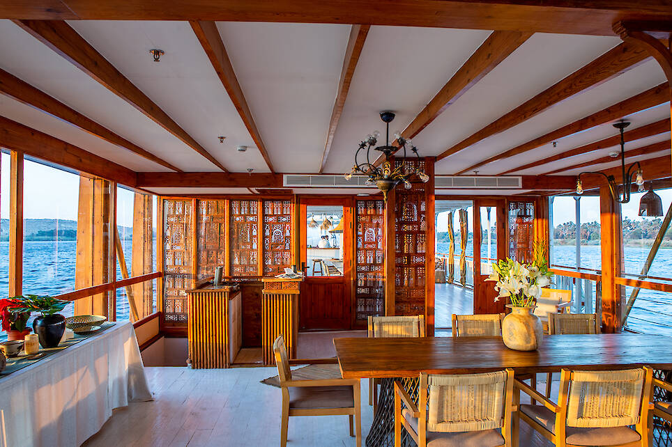 Interior of a wooden boat with large windows, a dining table with chairs, decorative flowers, and views of water and distant land through the windows