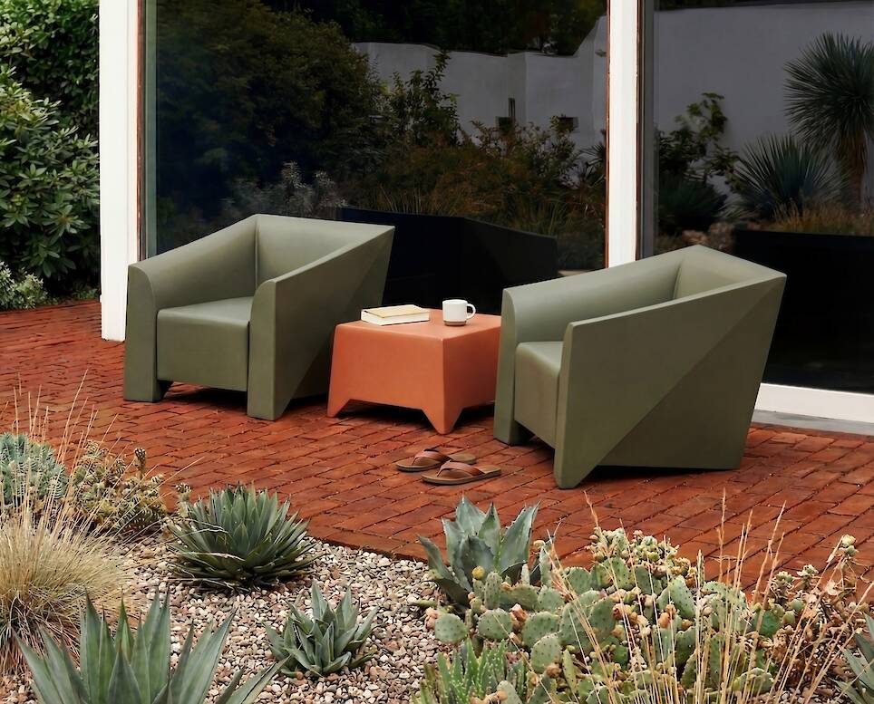 Two green armchairs and a small coffee table with a cup on it are arranged on a brick patio surrounded by desert plants