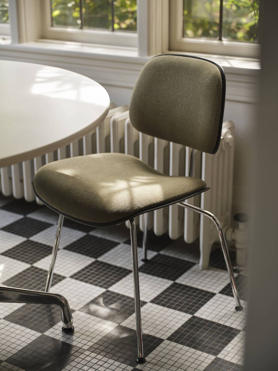 A green upholstered chair with metal legs sits next to a round white table on a black and white checkered tile floor by a window with sunlight streaming in