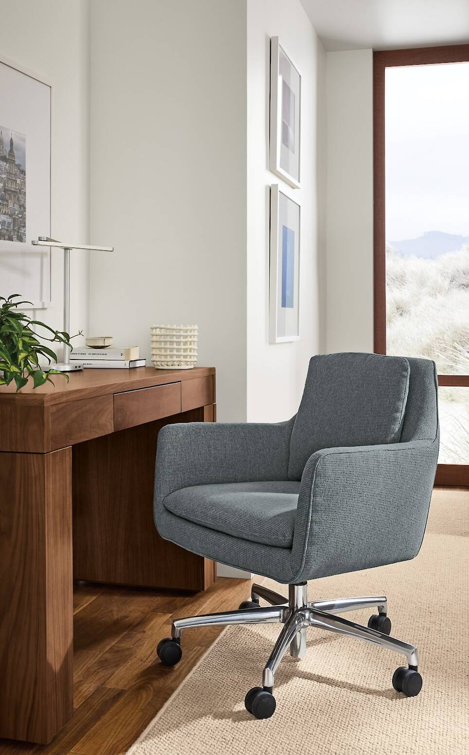 Gray office chair on wheels is positioned in front of a wooden desk, with framed pictures on the wall and a large window in the background