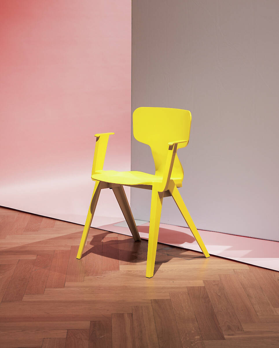 A modern yellow chair with angular legs and armrests sits on a wood floor against a background with pink and white gradients