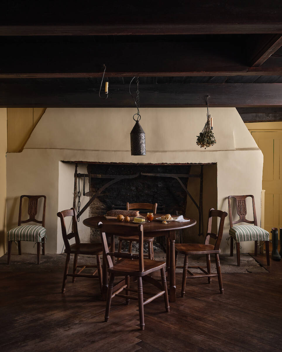 A rustic kitchen with a round wooden table, four chairs, food items on the table, and a large fireplace in the background