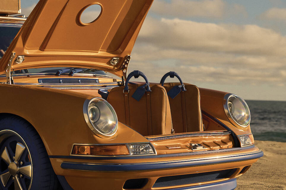 A vintage orange Singer Porsche 911 sports car with its front trunk open, showing two matching brown Louis Vuitton travel bags inside, parked by the ocean under a cloudy sky