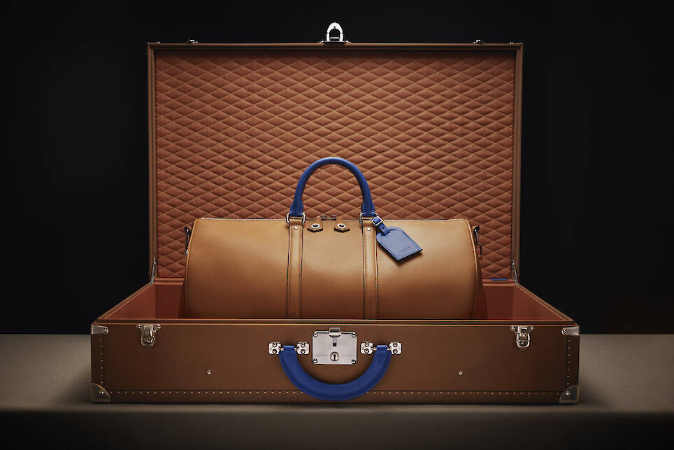 A brown leather duffel bag with blue handles sits inside an open, quilted brown suitcase against a dark background, evoking the timeless luxury of a Singer-restored Porsche 911