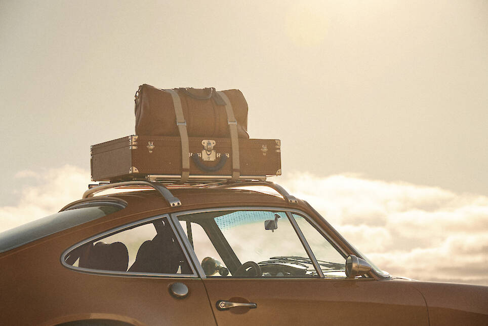 A brown vintage car, reminiscent of a Singer Porsche 911, with a roof rack carries a brown duffel bag and a hard suitcase against a bright sky—evoking the timeless elegance of