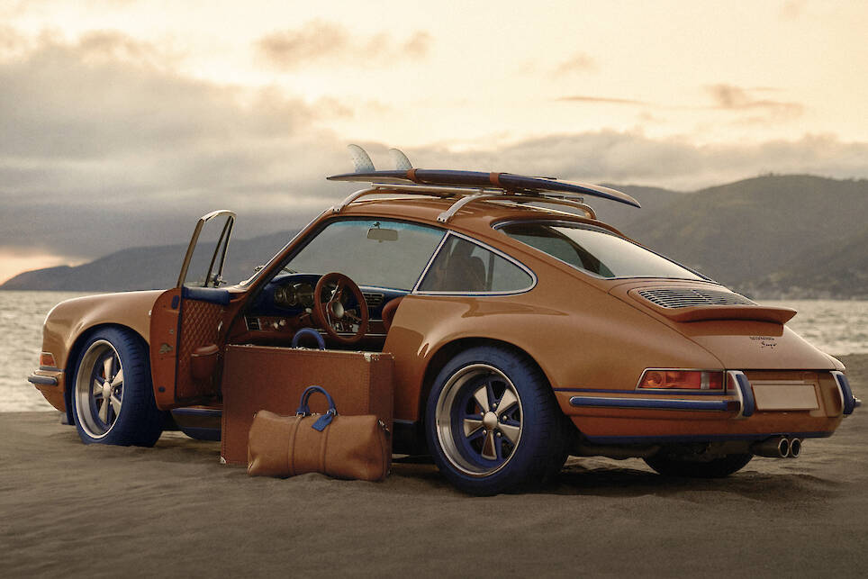 A vintage brown Singer Louis Vuitton Porsche 911 with a surfboard on the roof and two brown travel bags sits parked on a sandy beach at sunset near the ocean