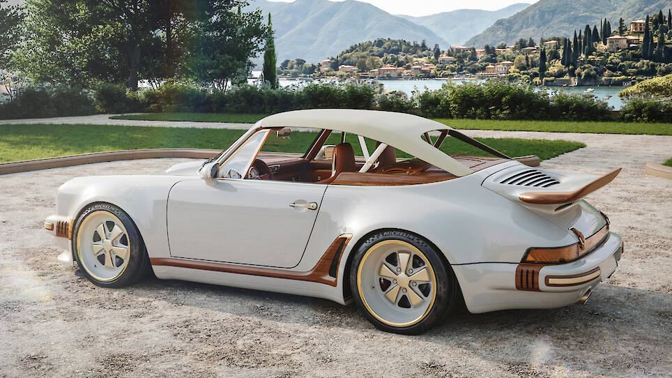 A white classic-style Singer Porsche 911 sports car with brown and gold accents is parked on a gravel path by a lake, with mountains and houses visible in the background