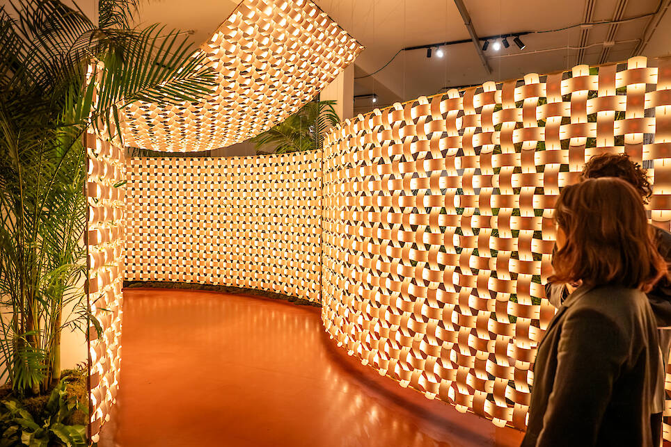 Two people observe a curved, illuminated wall installation—reminiscent of Rick Tegelaar’s innovative approach—made of stacked, glowing cylindrical elements in an indoor ex