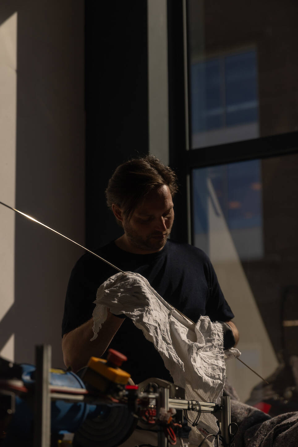 A person in a dark shirt uses a white cloth to clean or polish a long metal rod near a window, with sunlight casting shadows in the room—reminiscent of the meticulous craftsma
