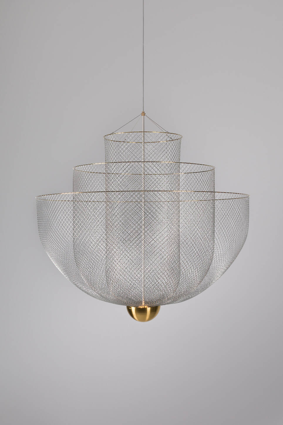 A tiered, wire mesh chandelier by Rick Tegelaar, featuring a gold spherical base, hangs elegantly from the ceiling against a plain gray background