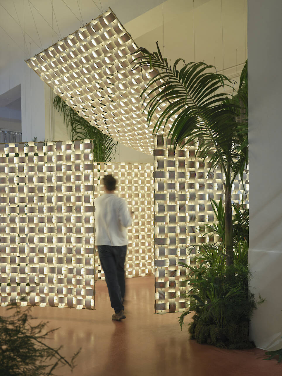 A person walks through an illuminated installation made of patterned light panels, designed by Rick Tegelaar, surrounded by indoor plants on a red floor