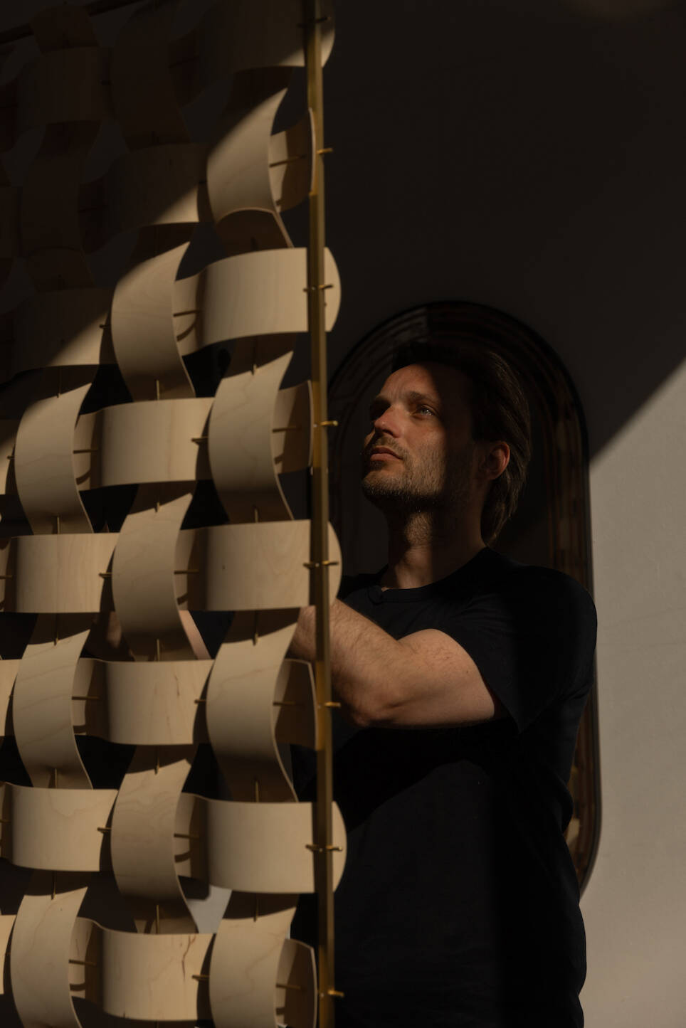 A man in a dark shirt stands next to a wooden art installation with curved pieces, illuminated by a beam of light in an otherwise shadowed room—a scene reminiscent of Rick Teg