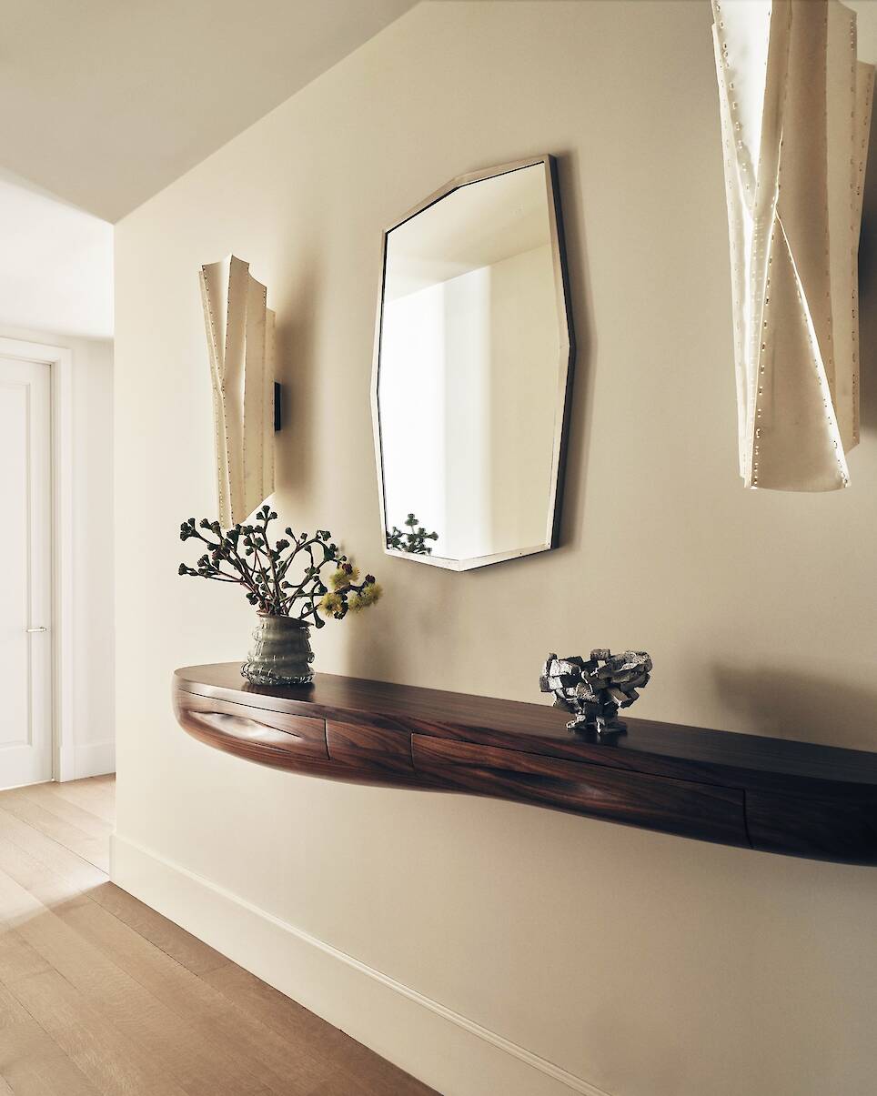 A hallway with a wooden floating console, a vase with branches, a decorative bowl, an octagonal wall mirror, and two sculptural wall sconces
