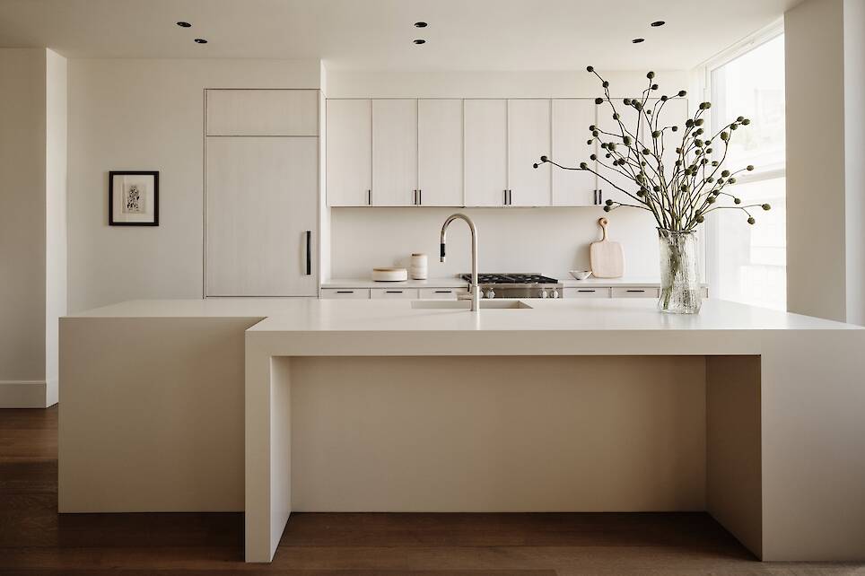 Minimalist kitchen with white cabinets, a large island with a built-in sink, wooden floors, and a glass vase with tall branches on the counter