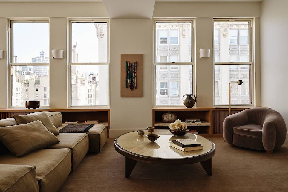 Minimalist living room with beige sofa, round coffee table, brown armchair, built-in shelves, and large windows showing a cityscape. Neutral tones and simple decor are featured