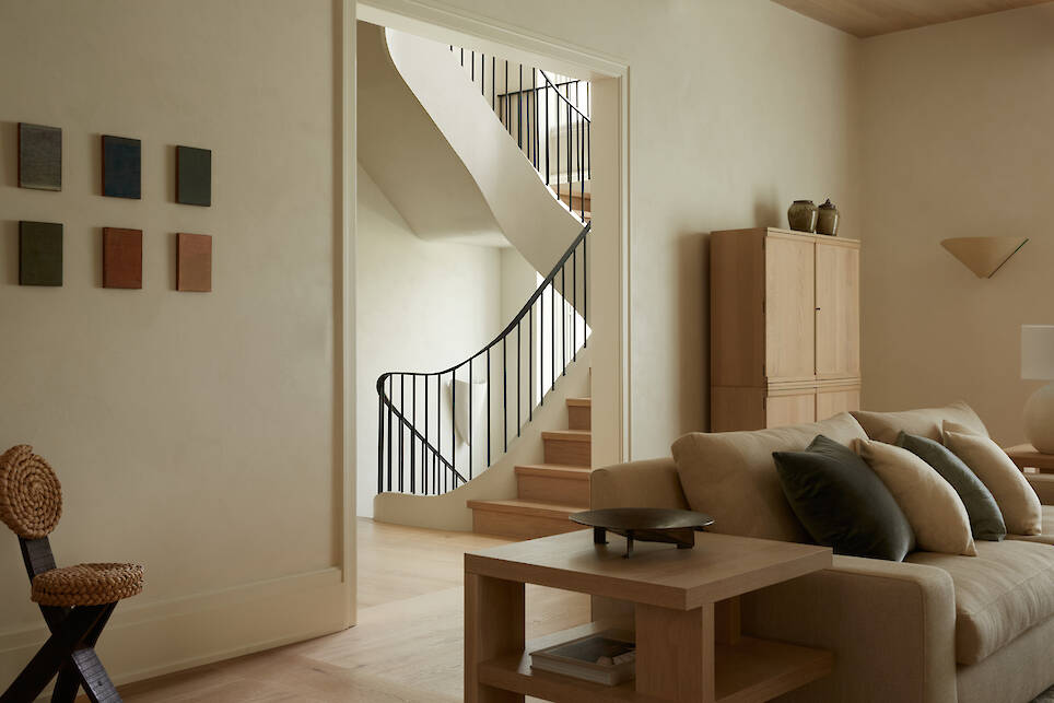 Modern living room with beige sofa, black and wooden accents, wall art, and a view of a staircase with a black railing in the background