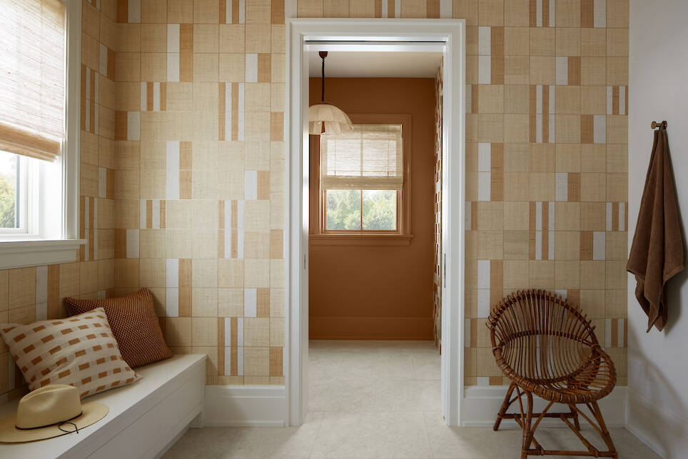 A hallway with patterned beige walls leads to a room with brown walls, wicker chair, window, and hanging light. Cushioned bench and towel hang beside the doorway