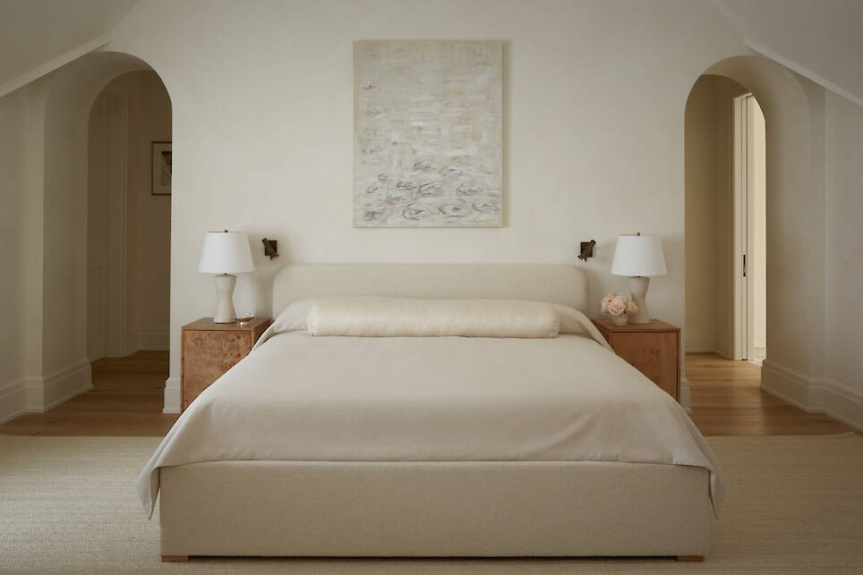 A minimalist bedroom with a beige bed, two wooden nightstands with white lamps, abstract art above the bed, and two arched doorways on either side