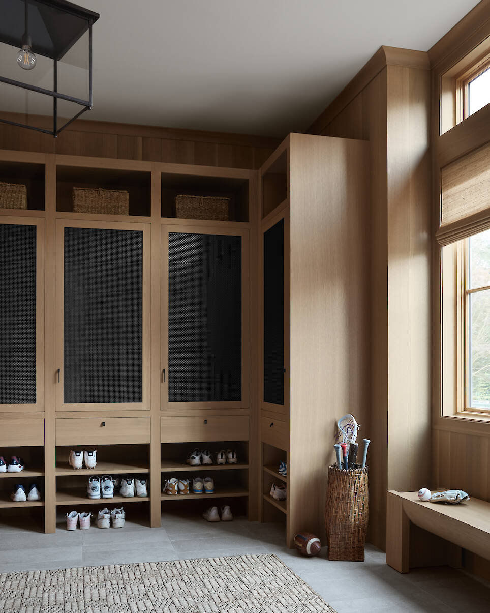 Mudroom with wooden lockers, shoe cubbies, a woven basket holding sports equipment, a window with a shade, and a bench beside a neutral patterned rug