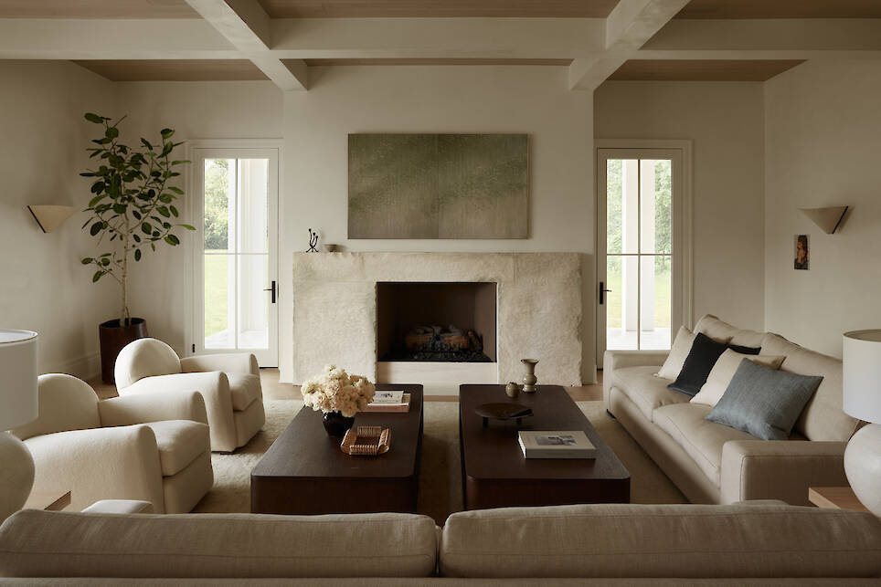 Neutral-toned living room with two sofas, two armchairs, a stone fireplace, wooden coffee tables, a potted plant, and minimal decor. Light streams in from two glass doors