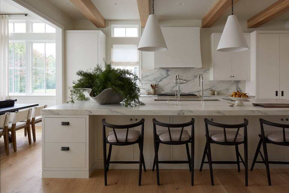 Modern kitchen with a marble island, three black chairs, pendant lights, white cabinets, and a dining area with large windows in the background