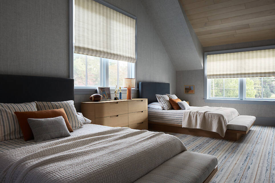 Bedroom with two beds, a wooden dresser between them, striped bedding, large windows with shades, and soft natural light