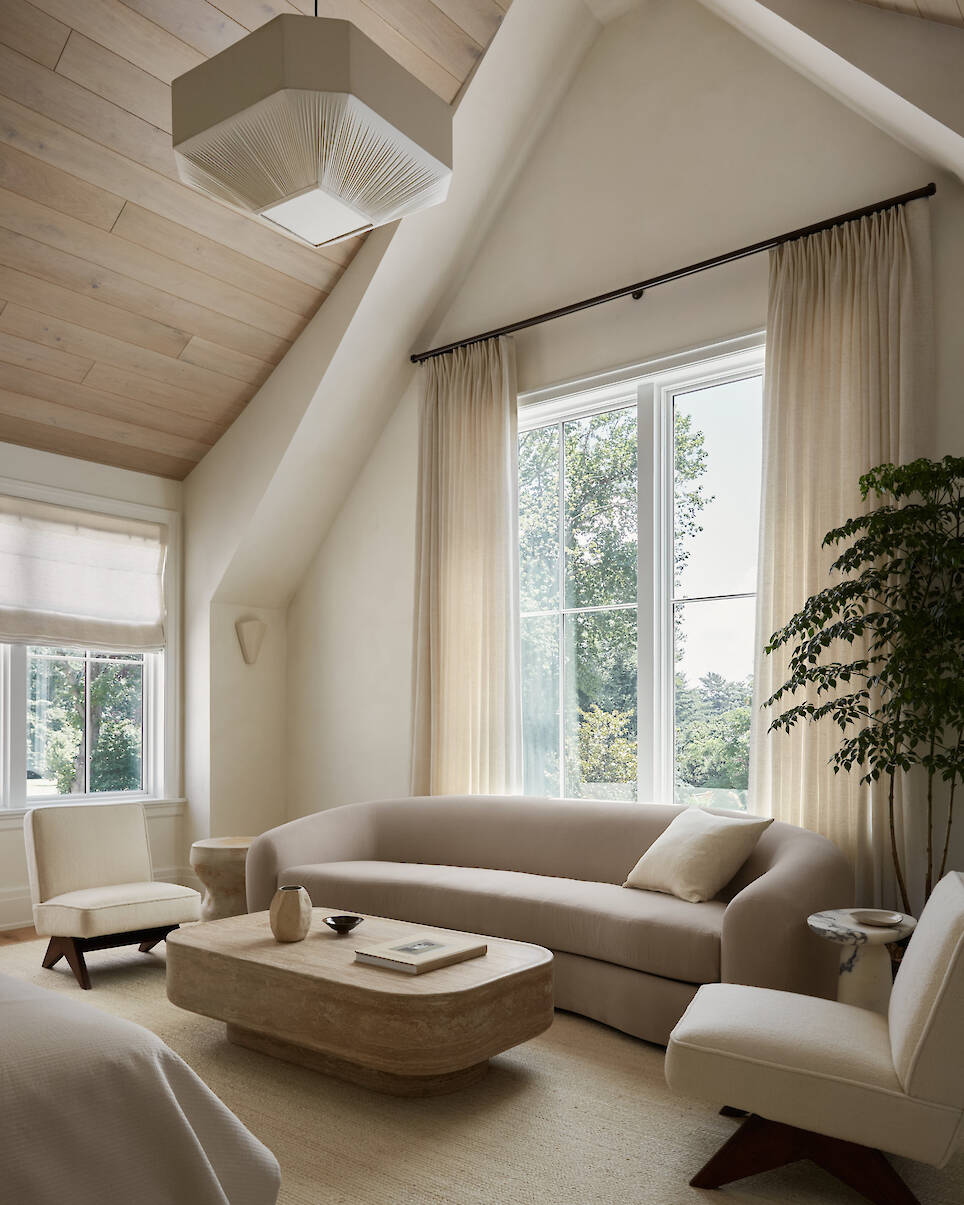 Neutral-toned living room with a curved sofa, wooden coffee table, two chairs, large windows, and a potted plant. Light streams in, highlighting minimalist decor and natural mat
