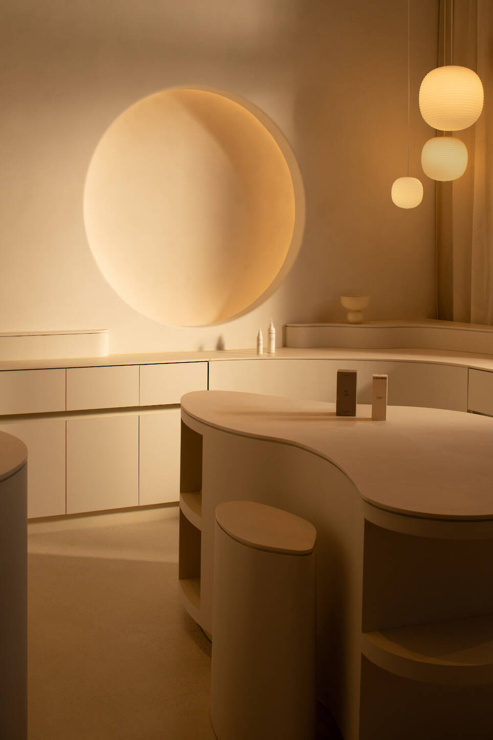 Minimalist beige room with curved counters, round wall feature, three hanging globe lights, and two bottles on the counter—capturing the serene elegance of Sana Skin Studio Wy