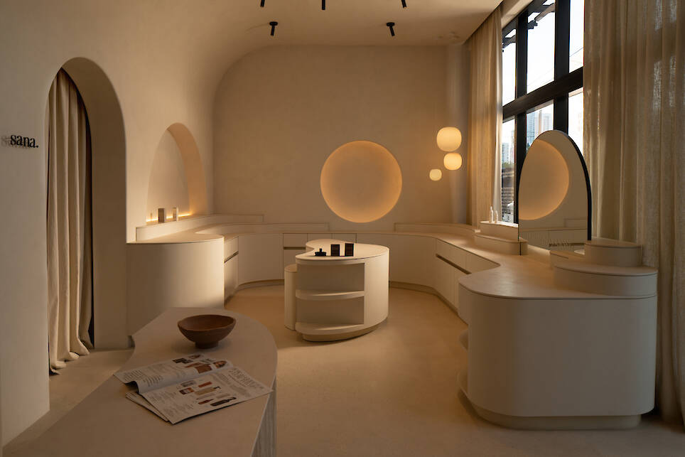 Minimalist beige room with curved counters, round wall lights, pendant lamps, large windows, curtains, a bowl, and a newspaper on display—reminiscent of the serene atmosphere