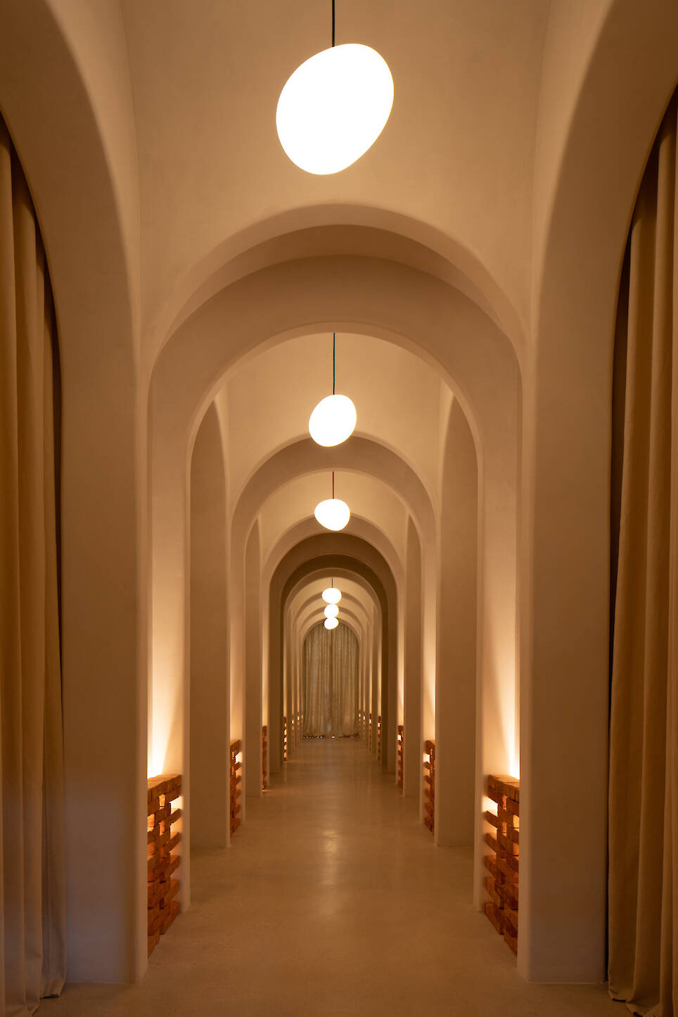 A long, arched hallway with beige walls, hanging round lights, soft floor lighting, and tan curtains on both sides creates a serene ambiance reminiscent of Sana Skin Studio Wynw
