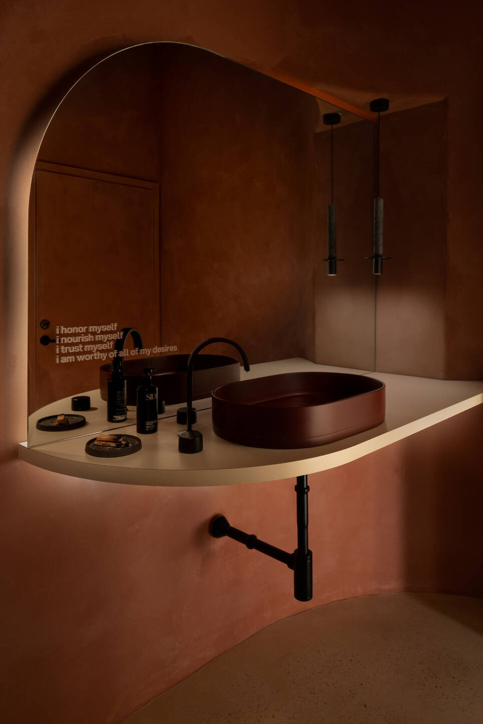 Modern bathroom with curved beige countertop, maroon oval sink, matte black fixtures, toiletries, and affirmations on a brown textured wall—evoking the calming atmosphere of S