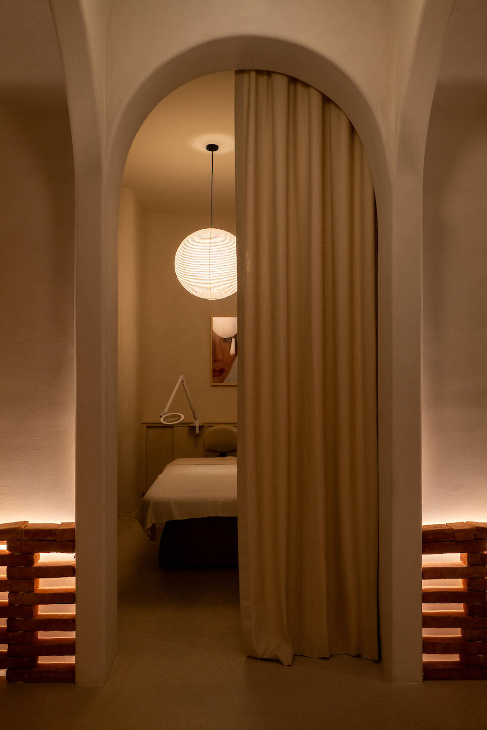 A softly lit treatment room at Sana Skin Studio Wynwood features a massage table, a round paper lantern, a curtain partition, and minimalist decor in neutral tones