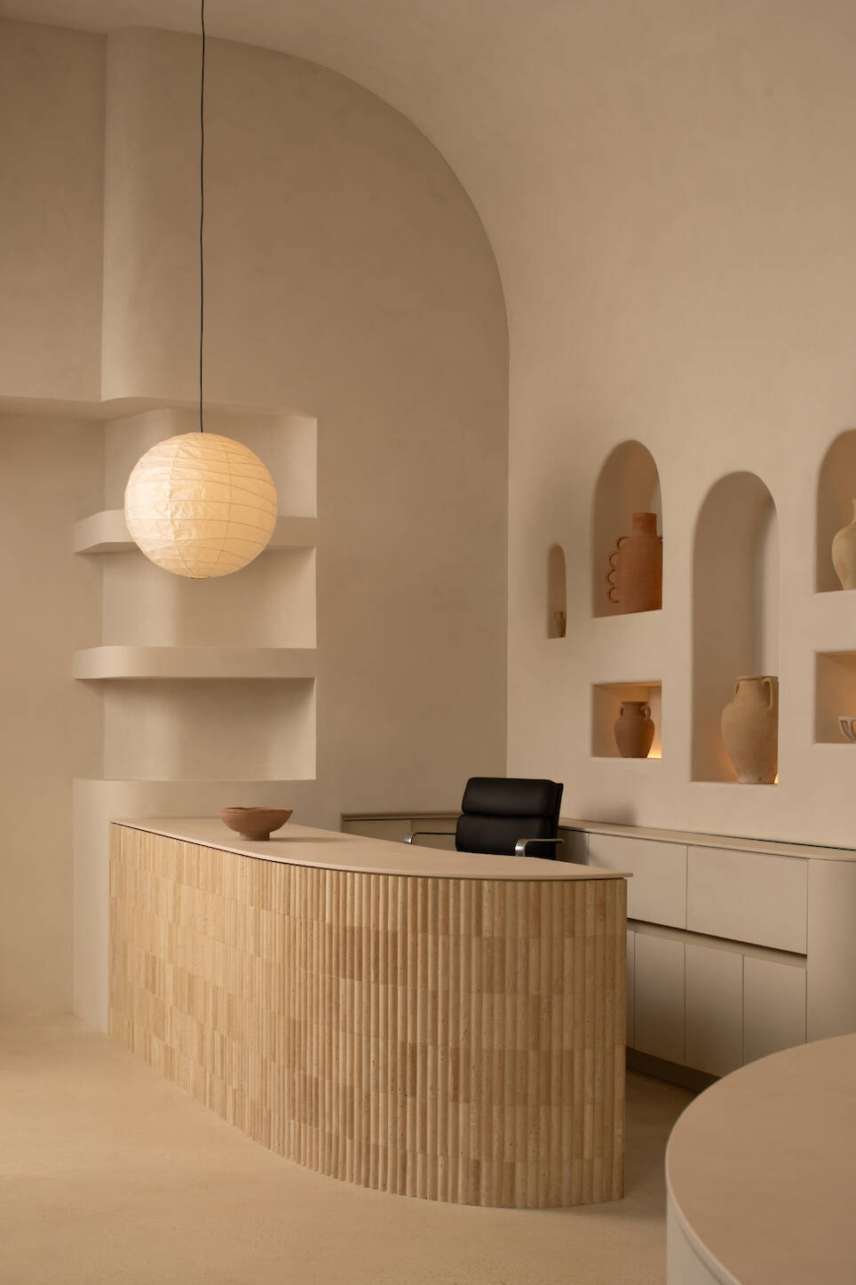 Curved wooden reception desk with a black chair, built-in shelves, and pottery accents in a beige minimalist room at Sana Skin Studio Wynwood, beautifully lit by a round pendant