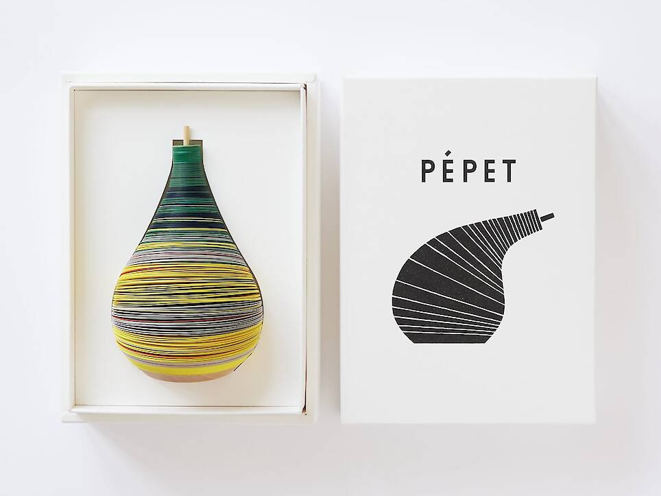 a small paper figure made from numerous layers of colored paper on a flexible central axis so that it looks like an anthropomorphized pear, nestled inside of a box