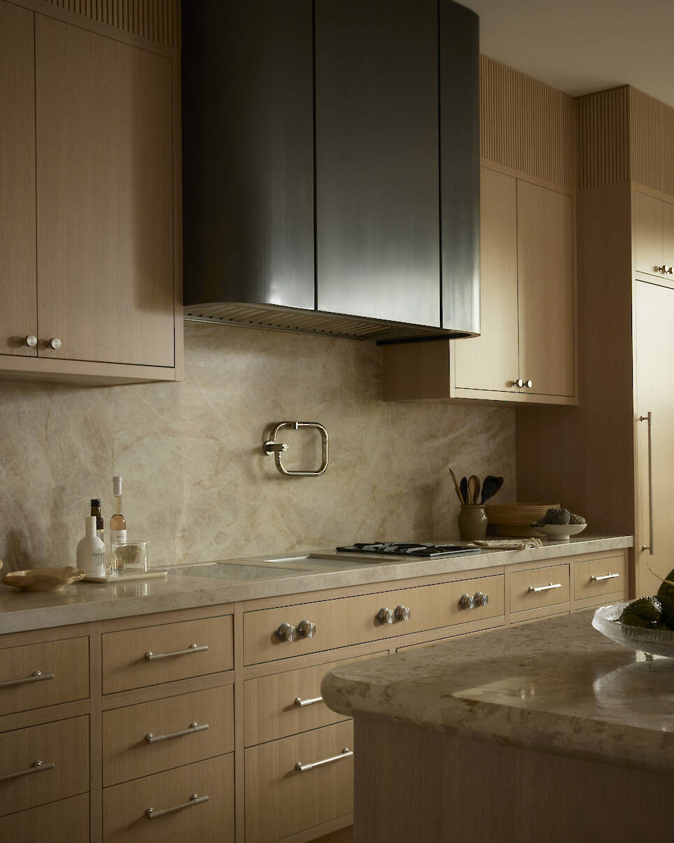 Modern kitchen with light wood cabinets, marble countertops, a marble backsplash, stainless steel gas stove, black range hood, and minimal accessories on counters
